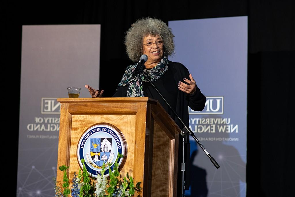 Angela Davis stands at a podium in front of a screen displaying the U N E logo