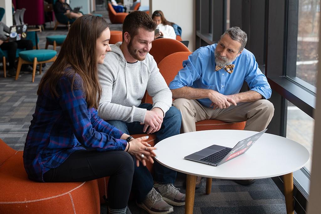 A U N E professors works with two U N E liberal arts students at a table with a lap top