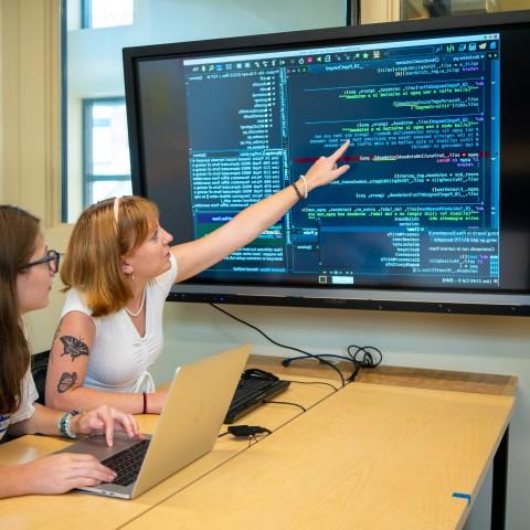 Two female students sit in front of a computer with large numbers of data appearing on a TV screen